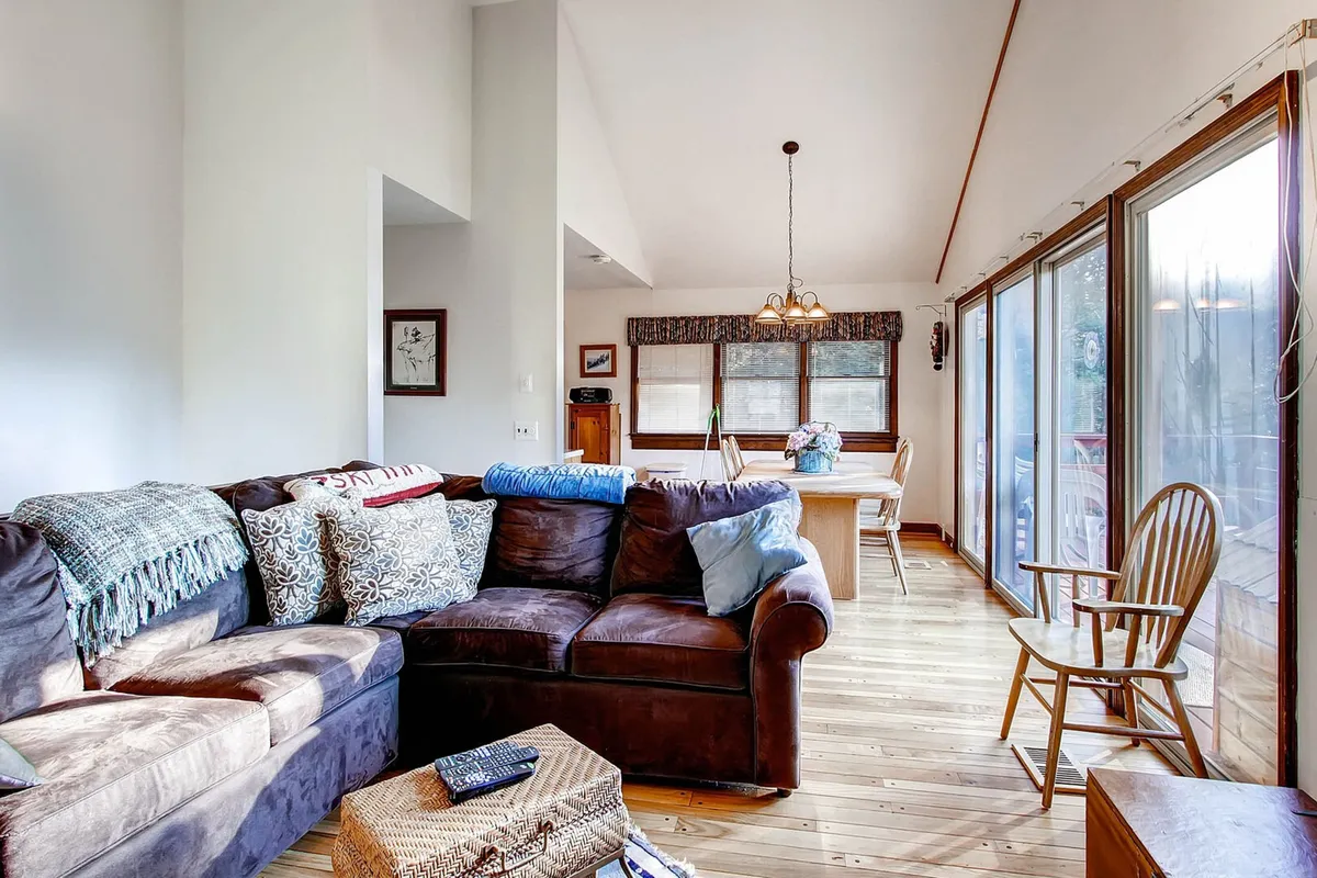 A spacious, light-filled living and dining area features a large sectional sofa by broad windows, offering inviting views outdoors. The space exudes warmth and provides a comfortable spot to relax after a day outside.