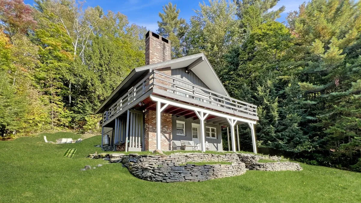 The exterior of the chalet, featuring a rustic house structure with a stone wall, inviting porch, and surrounding trees. The scene is cozy and welcoming, suggesting a perfect home base to return to after a day on the mountain and a great place for group gatherings or relaxing outdoors.