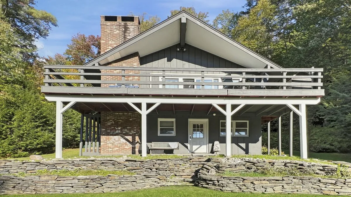 The hero image shows the Vermont chalet’s striking exterior with its unique roofline, expansive balcony, and inviting stone wall, set against a backdrop of lush mountain scenery. The spacious balcony is perfect for gatherings, offering panoramic sunset views and plenty of space for morning coffee or evening barbecues with family and friends. The overall mood is warm, welcoming, and nestled in nature.