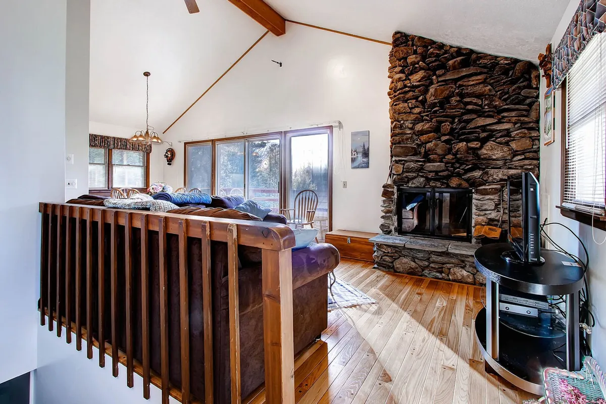 A cozy living room featuring a natural stone fireplace flanked by large windows, a plush sofa positioned to take in both the crackling fire and the scenic views outside, and warm wooden accents that lend a welcoming, rustic atmosphere.