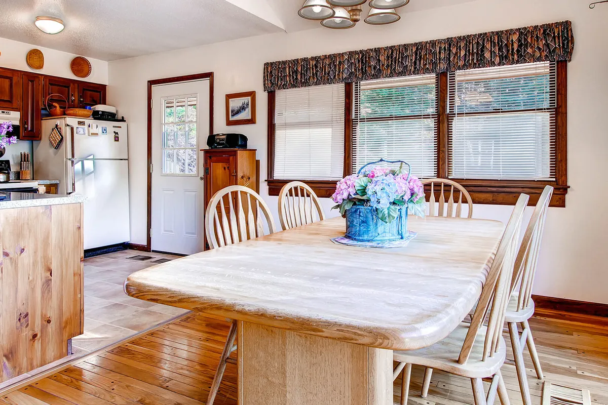 A bright, inviting dining area with a large wooden table set for a meal, a floral centerpiece, ample seating, and kitchen cabinetry visible in the background. Sunlight bathes the room, emphasizing its role as a communal space.