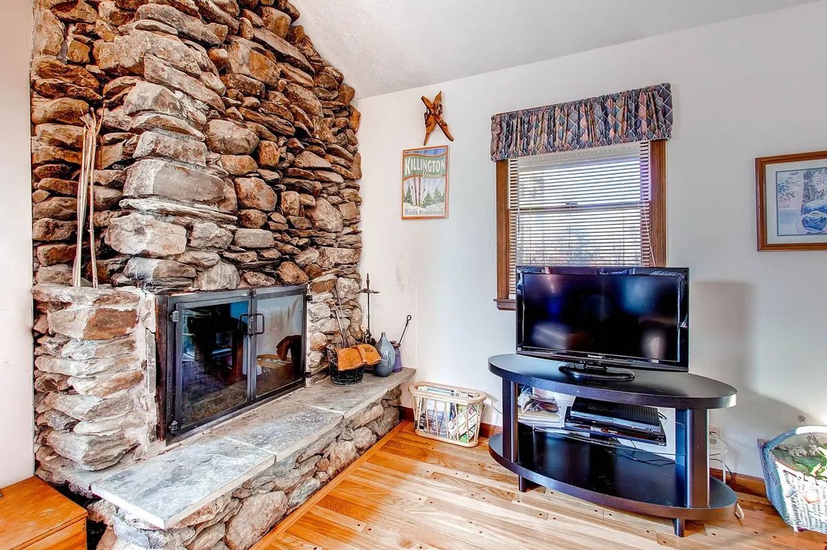 A living space centered around a beautiful stone fireplace, with a comfortable couch and wooden flooring. Large windows suggest views of the outdoors, inviting guests to relax, warm up, and share stories after outdoor adventures.