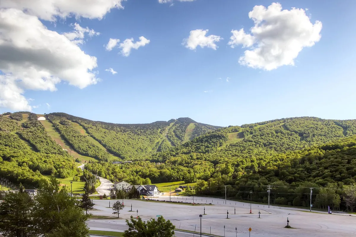 This image captures sweeping views of Vermont mountains framed by lush greenery under a clear blue sky. The scene includes a vacant parking lot, suggesting easy access to outdoor activities and trails.